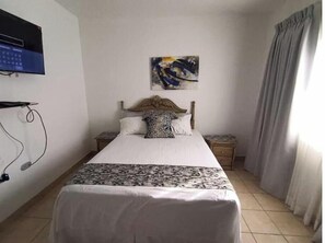A bedroom with a bed, a TV mounted on the wall, a painting above the bed, and a window with curtains.