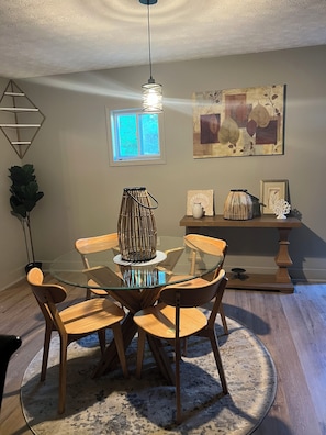 Un comedor con mesa de vidrio, sillas de madera, lámpara colgante, ventana y decoración en la pared.