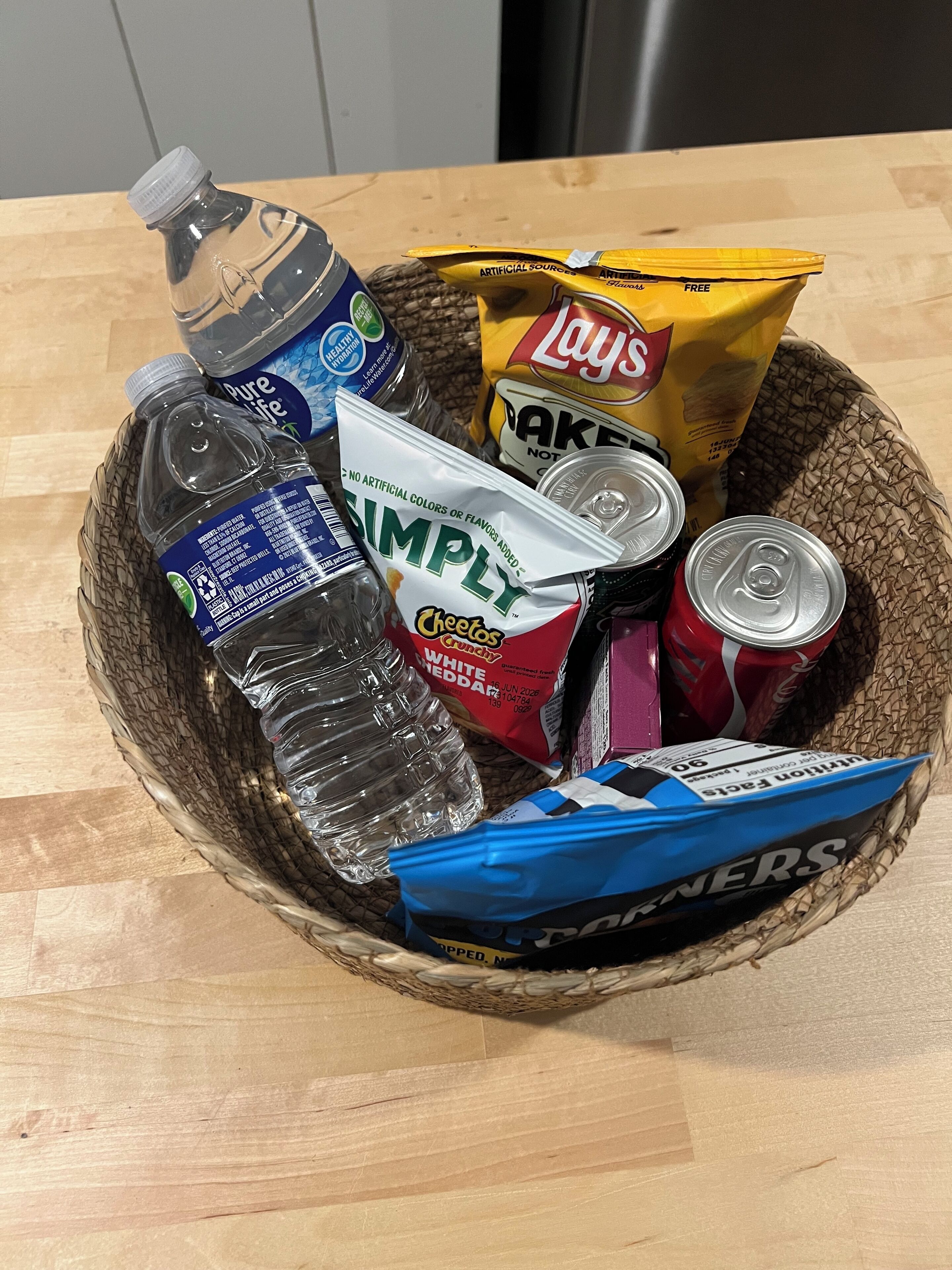 A basket containing a bottle of water, a bag of Lay's chips, a can of soda, a bag of Simply white cheddar cheese, and a packet of instant noodles.