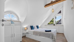 Ett sovrum med ett sluttande tak, en säng med ett randigt överkast, en vit garderob, ett litet skrivbord med en lampa och ett fönster med utsikt över omgivningen.