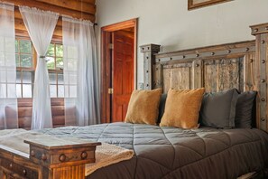 A rustic bedroom with a wooden headboard, a bed with a quilted blanket, and a window with sheer curtains. - Cozy Rustic Cabin Close to Town w/Hot Tub & Games (Sevierville)