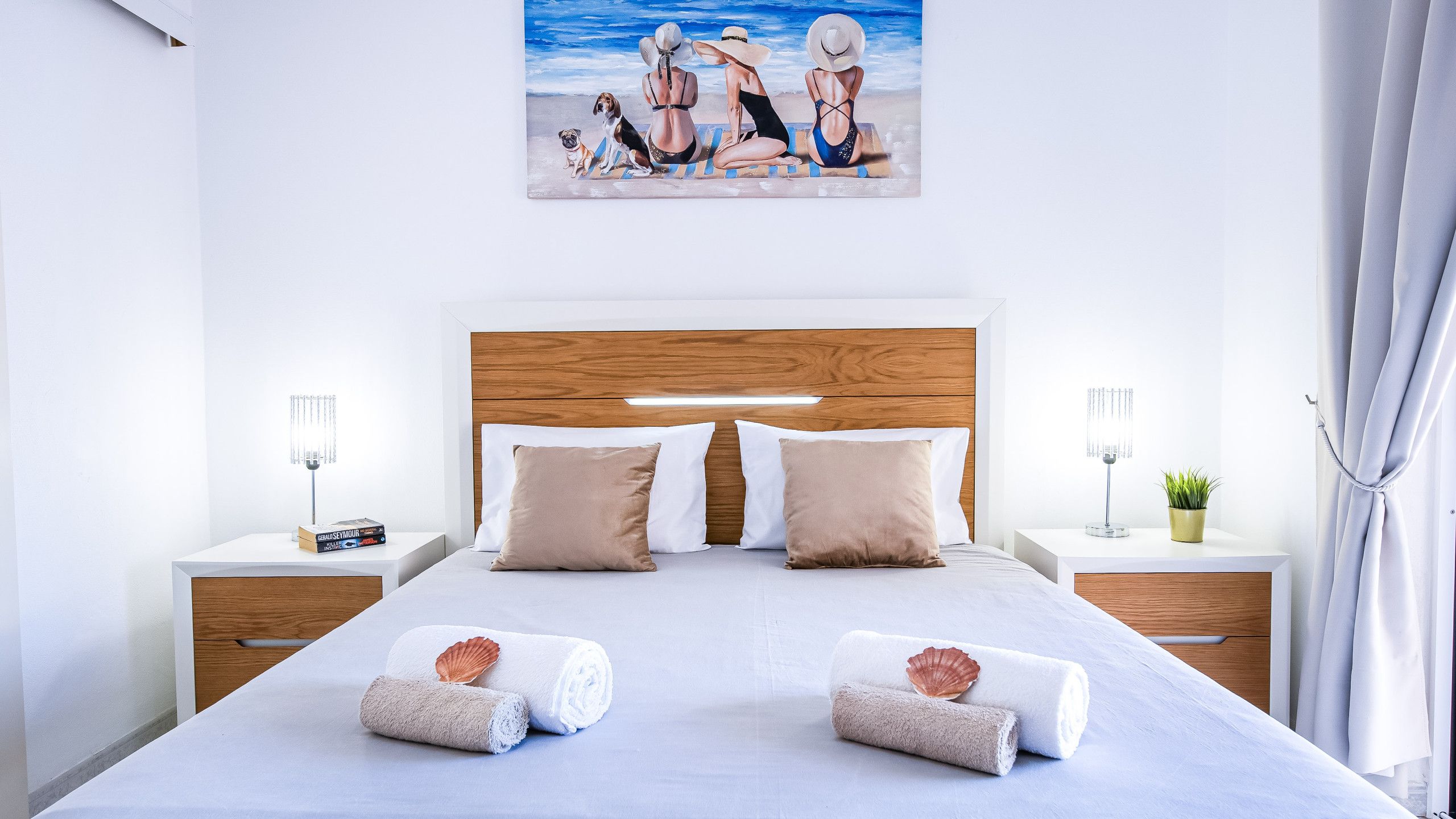 A neatly made bed with two pillows and two rolled towels, a wooden headboard, bedside tables with lamps, and a painting above the bed.
