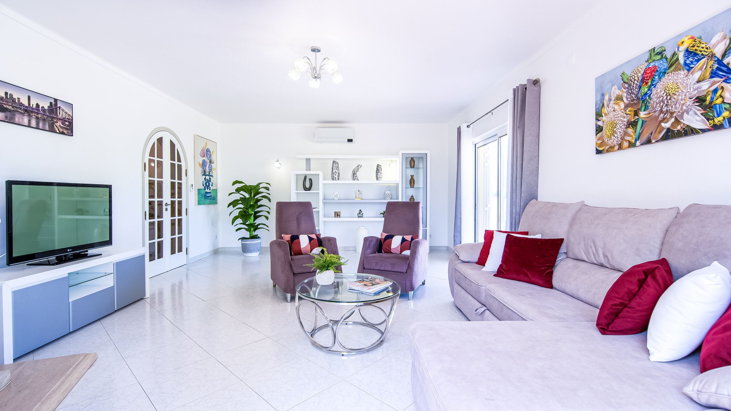 Een moderne woonkamer met een flatscreen televisie, een bank met rode en witte kussens, een glazen salontafel en een potplant.