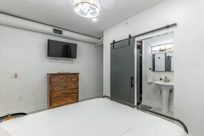 A bedroom with a bed, a wooden dresser, a television, a sliding door, and a bathroom with a sink and mirror. - Downtown Chattanooga Condo: Modern Suite w/ City (Chattanooga)