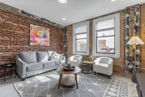 Una sala de estar con pared de ladrillo, un sofá gris, sillones blancos, una mesa de centro redonda y vistas de edificios a través de grandes ventanales.