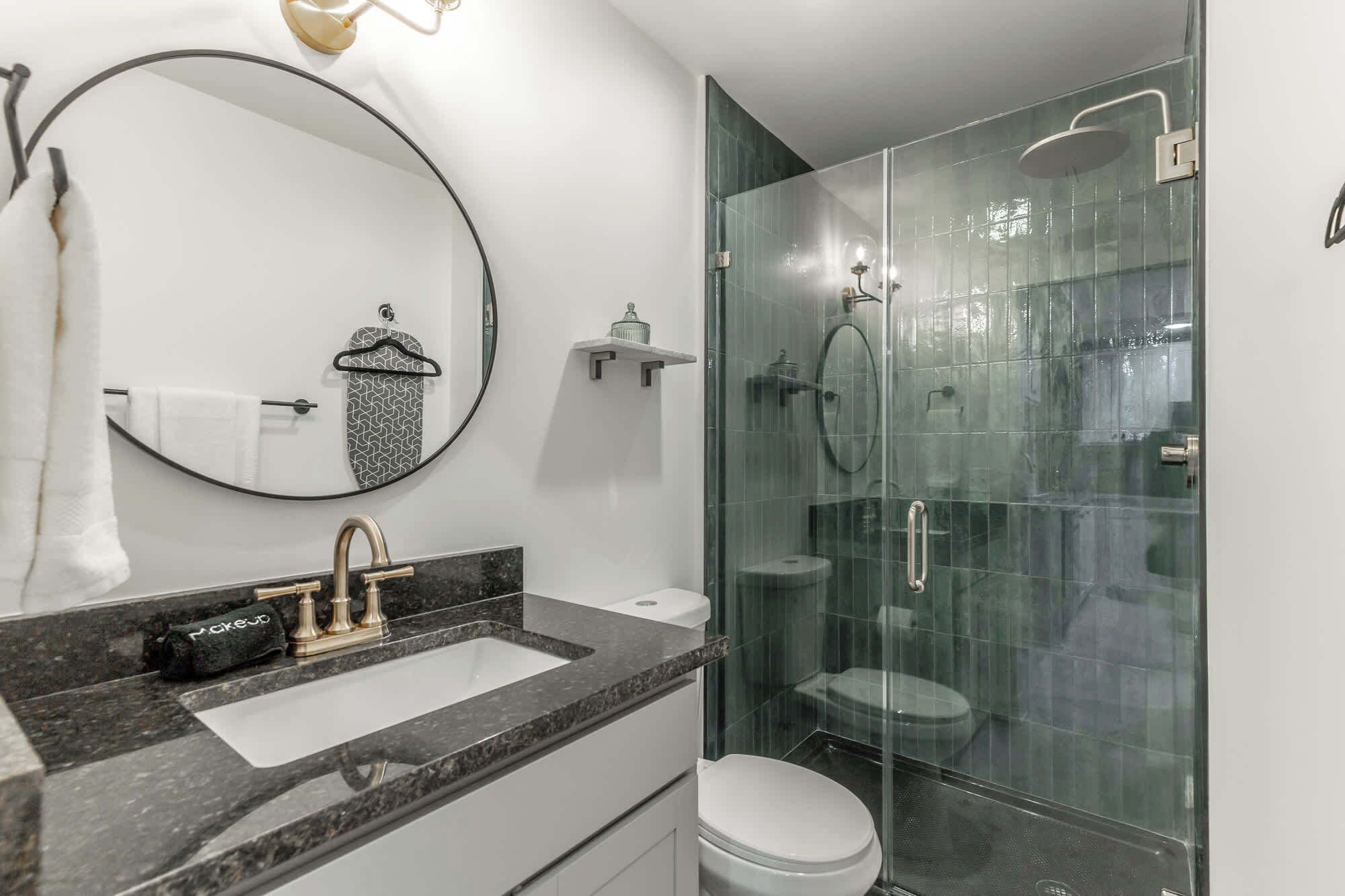 A modern bathroom with a round mirror, a sink with a gold faucet, a toilet, and a glass-enclosed shower.