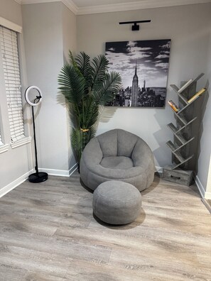 Ein Zimmer mit grauem Sitzsack, einem kleinen Hocker, einer hohen Stehlampe, einem Topfpflanzen, einem gerahmten Stadtbild und einem Holzregal mit Büchern.