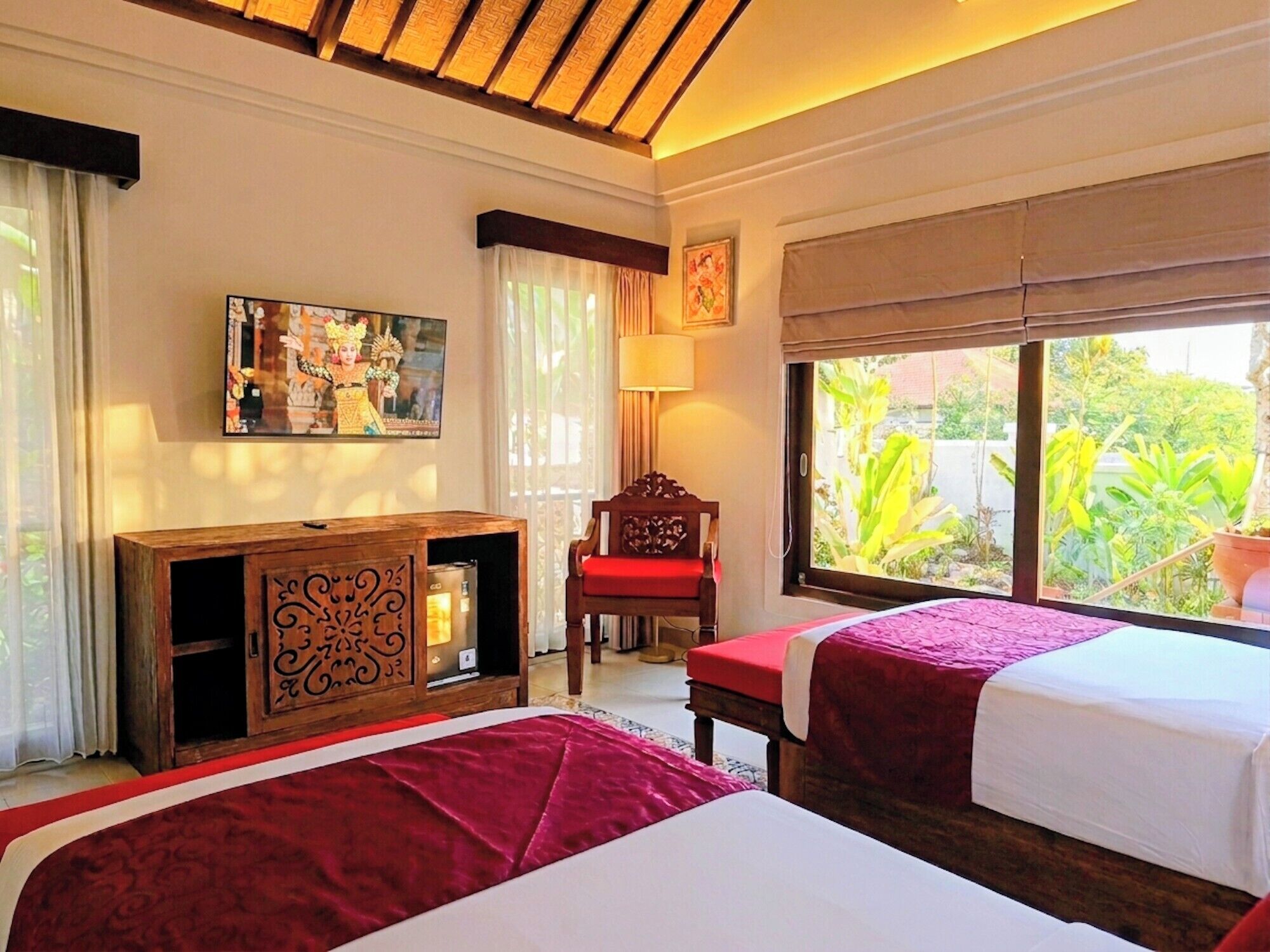 A hotel room with two beds, a wooden cabinet, a chair, a window with a view of greenery, and a painting on the wall.