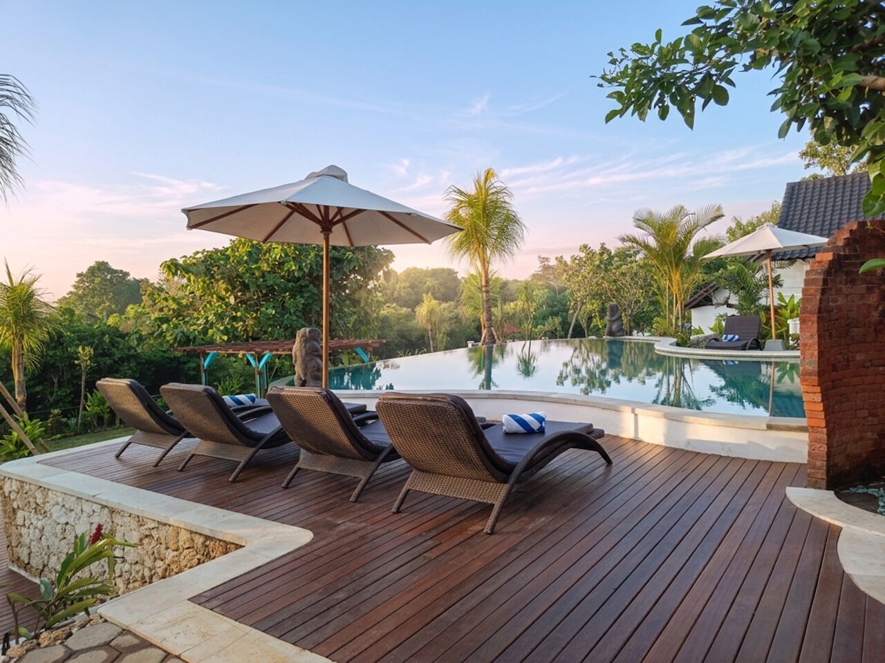 A wooden deck with lounge chairs and umbrellas, a swimming pool, and lush greenery.