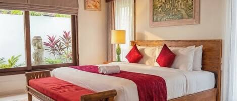 Un dormitorio con cama de madera, colcha roja, alfombra estampada, ventana con vista a follaje y techo con vigas de madera.