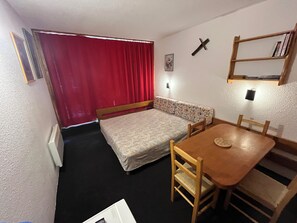 Une petite pièce comprenant un lit, une petite table et une chaise. On y trouve un rideau rouge, une étagère fixée au mur avec des livres, et une décoration en forme de croix.