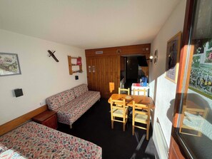 Une pièce de taille modeste comprenant un canapé, une petite table avec des chaises, un meuble en bois, une étagère fixée au mur et une porte donnant sur une autre pièce.