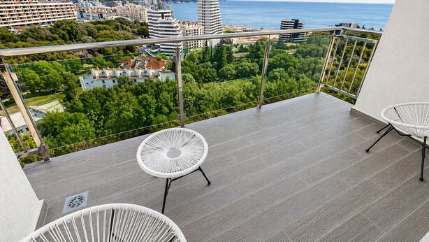 Un balcon avec deux fauteuils en osier blancs, un sol carrelé gris et une vue sur le paysage urbain et la mer.