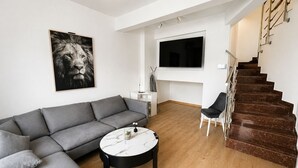 Un salon moderne avec un canapé d’angle gris, une table basse ronde, un téléviseur à écran plat fixé au mur et un escalier en bois.