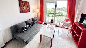 A modern living room with a grey sofa, a white coffee table, a red TV stand, and a large window with red curtains. - Beachfront duplex - La Arena 4608 (Isla)