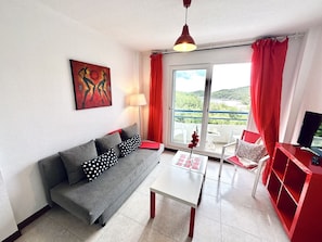 Una sala de estar moderna con un sofá gris, una mesa de centro blanca, un mueble de televisión rojo y un ventanal con cortinas rojas.