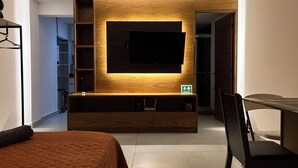 Een moderne hotelkamer met een grote televisie bevestigd aan een houten muur, een bed met een bruine spreidek, een eettafel met stoelen en een kleine kitchenette.