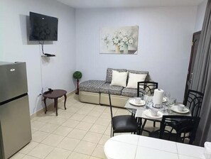 A compact living space with a kitchenette, a sofa, a dining table with chairs, and a flat-screen TV mounted on the wall.