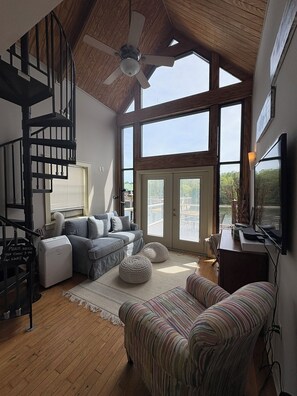 A spacious living room with a high ceiling, a built-in fireplace, a large window, and a staircase.