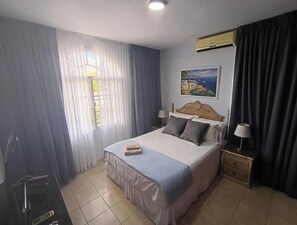 A hotel room with a bed, a nightstand with a lamp, a painting on the wall, and a window with curtains.