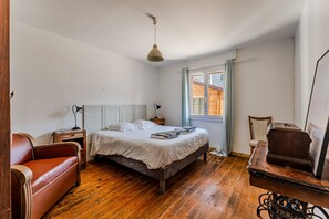 A bedroom with a wooden floor, a bed with a white bedspread, a brown leather sofa, a wooden nightstand, a desk with a chair, and a hanging light fixture. - Holiday Home “Enjoy the Spa All Year Round” with Private Terrace, Garden & Wi-Fi (La Teste-de-Buch)