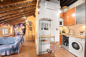 Una cocina compacta con lavadora, un estante metálico con utensilios y una cama con ropa de cama estampada.
