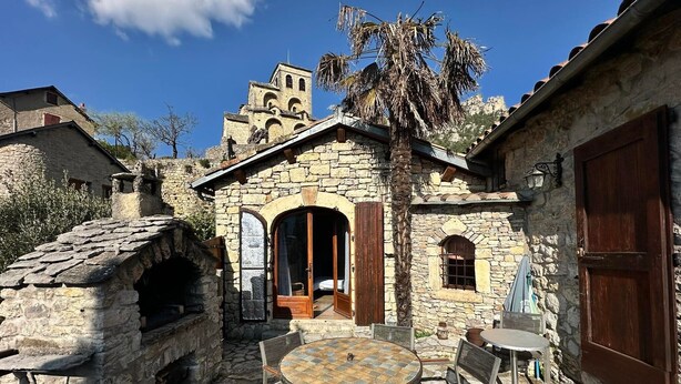 Un edificio de piedra con una mesa redonda y sillas, una puerta de madera y vistas a una montaña.