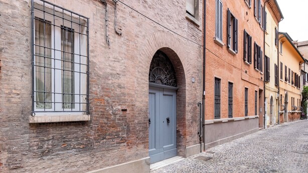 A cobblestone street with a brick building featuring a blue door and window with metal bars. - Segreto Estense: Dormi a un Passo dalla Storia (Ferrara)