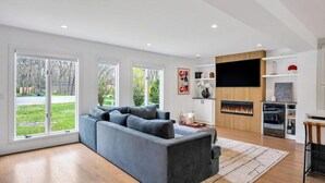 Un salon moderne avec un canapé d’angle gris, un téléviseur à écran plat, une cheminée et une vue sur l’extérieur.