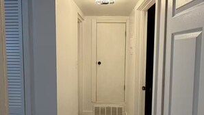 A hallway with a white door at the end, a vent on the wall, and a ceiling light fixture.