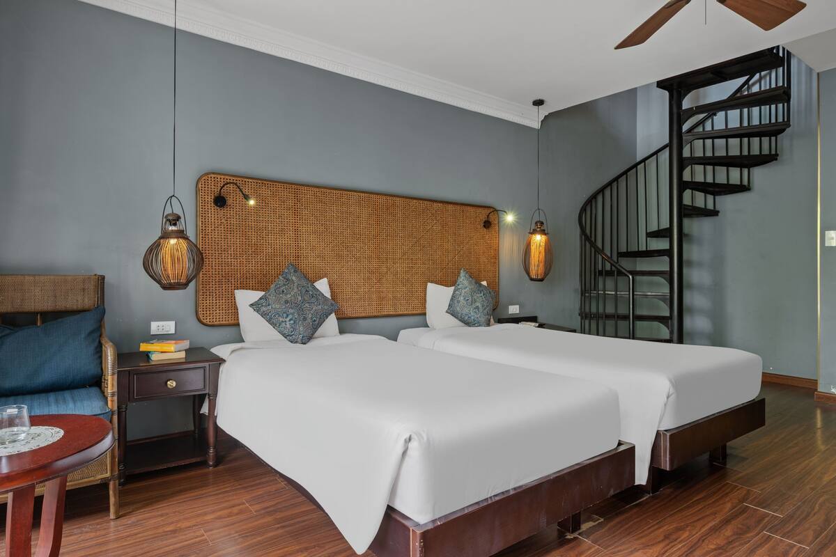 Habitación de hotel con dos camas, cabecero de ratán, piso de madera, escalera en espiral y una pequeña mesa con un vaso.