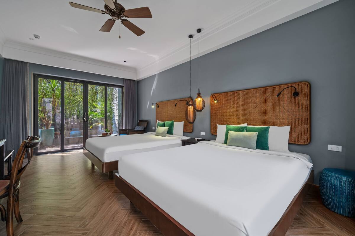 Habitación de hotel con dos camas, ventilador de techo, ventana grande y zona de comedor.