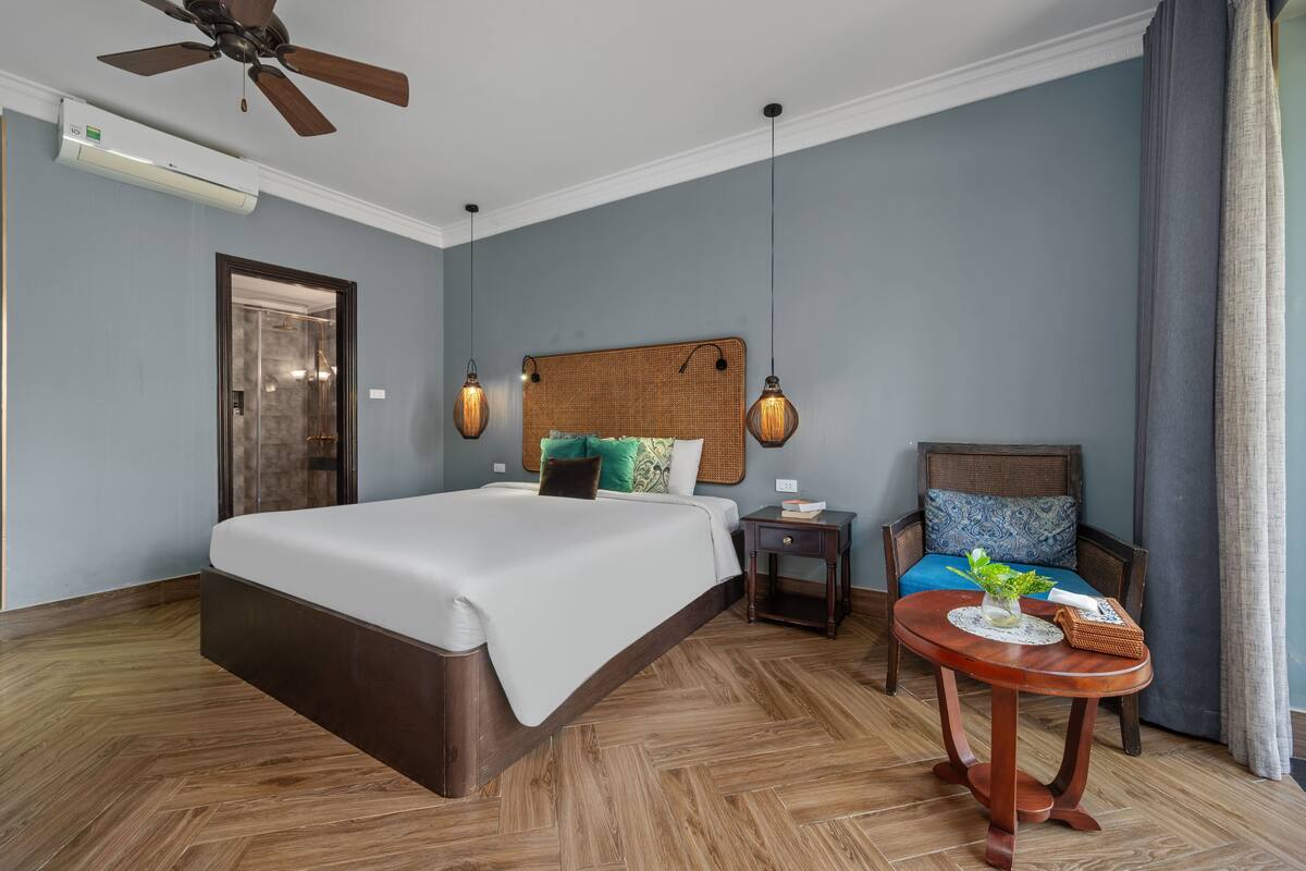 Habitación de hotel con cama, una silla, una mesita, ventilador de techo y aire acondicionado instalado en la pared.