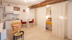 Un espacio habitable compacto con kitchenette, un área de comedor con sillas rojas, una cama con cortinas blancas y una viga de techo de madera.