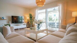 A living room with a sofa, a coffee table, a TV, and a large window with curtains. - Happy Hideaway - Private Resort Week, Sleeps 6 (Jan Thiel)