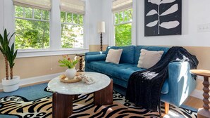Ein Zimmer mit einem blauen Sofa, einem runden Couchtisch, einem zebra-gemusterten Teppich und einem großen Fenster mit Blick auf Bäume.