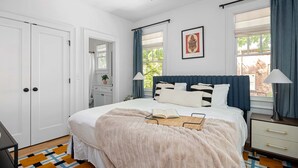 Ein Schlafzimmer mit Bett, Nachttischen, Deckenventilator, einem Fenster mit Vorhängen und einer Tür.