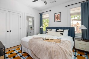 A bedroom with a bed, bedside tables, a ceiling fan, a window with curtains, and a door.