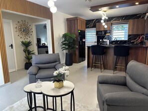 Una sala moderna con un sofá gris, dos mesas redondas, una planta en maceta y una zona de cocina con barra y taburetes.