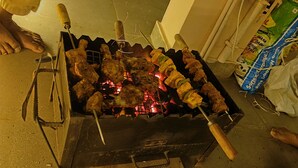 A grill with skewers of meat and vegetables, a person grilling, and a bag of groceries in the background.
