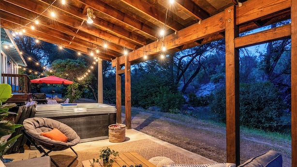 An outdoor patio with a hot tub, wooden furniture, string lights, and a view of the surrounding forest. - Hot Tub-Creekside Yosemite Family Oasis - 5 min to Town-Foosball, Pets Welcome (Mariposa)