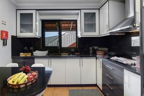 A kitchen with white cabinets, a black countertop, and a window with a view of stairs. - The Cozy Pink House – Ponta do Sol (Canhas)