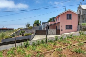A modern house with a sloped roof, a stone retaining wall, and a paved driveway. - The Cozy Pink House – Ponta do Sol (Canhas)