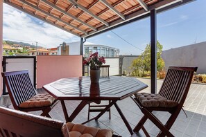 Un patio al aire libre con una mesa de madera y dos sillas acolchadas, una planta en maceta y vistas de edificios al fondo.