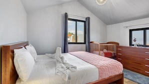 Ein Schlafzimmer mit Bett, Holz-Kopfteil, Kommode, Babybett und einem Fenster mit Vorhängen.