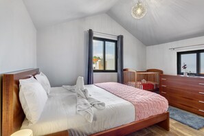 Un dormitorio con cama, cabecero de madera, cómoda, cuna y ventana con cortinas.