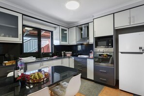 A modern kitchen with a black countertop, white cabinets, stainless steel appliances, and a window providing natural light. - The Cozy Pink House – Ponta do Sol (Canhas)