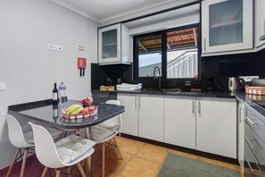 Una cocina con armarios blancos, encimera negra, ventana con vista a un edificio y zona de comedor con sillas blancas.