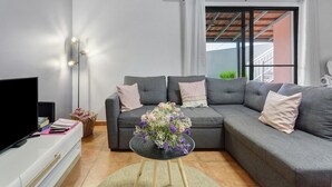 Ein Wohnzimmer mit einer Eckcouch, einem Flachbildfernseher, einem kleinen runden Tisch mit Blumenvase und einer Stehlampe.