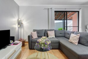 A modern living room with a sectional sofa, a flat-screen TV, and a small round table with a floral arrangement. - The Cozy Pink House – Ponta do Sol (Canhas)